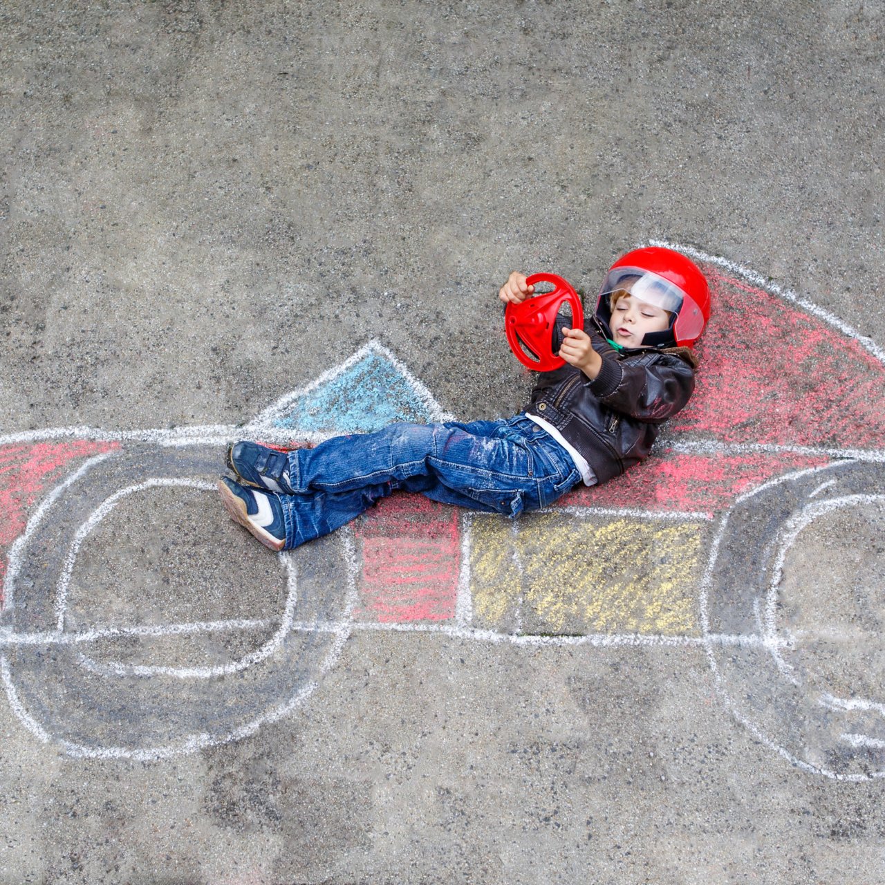 Auf dem Boden wurde ein Rennwagen aus Kreide gemalt, darauf liegt ein Junge mit einem Helm auf und einem Lenkrad in der Hand. 