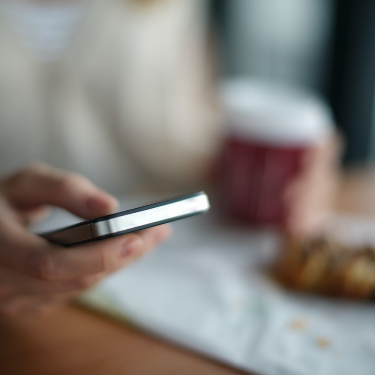 Person mit Handy in der Hand sitzt an einem Tisch und trinkt Kaffee. 